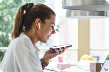 Woman with a ponytail sitting at her kitchen table on her laptop and taking a call with her wireless headphones.