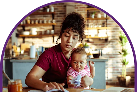 Curly-haired mother holds her child while doing bills at the kitchen table.