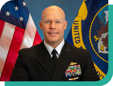 Official portrait of a U.S. Navy captain in dress uniform seated in front of the American flag and Navy flag