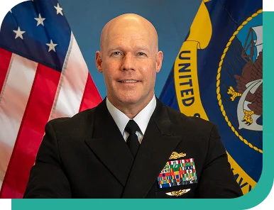 Official portrait of a U.S. Navy captain in dress uniform seated in front of the American flag and Navy flag