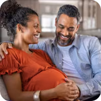 A pregnant brown-skinned woman in a dark orange blouse smiles as a smiling dark skinned man in a grey shirt rests a hand on her stomach.