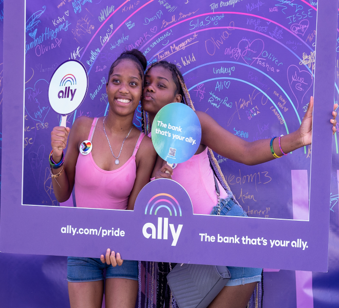 Two happy young women wearing matching pink tops pose with the Ally Pride frame and fans in front of the Allies Arc project.