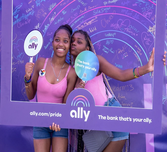Two happy young women wearing matching pink tops pose with the Ally Pride frame and fans in front of the Allies Arc project.
