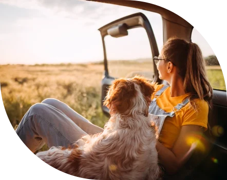 Image of a person with glasses and a ponytail wearing a yellow t-shirt and overalls sitting inside a vehicle next to a dog, both looking outside from the open door