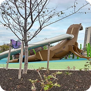 Newly planted trees and landscaping at Ralph C. Wilson, Jr. Centennial Park with a wooden dinosaur play structure in the background