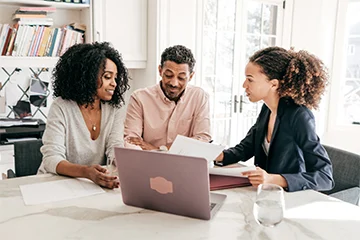 A young man and woman sit at a kitchen table with an advisor and laptop looking at paperwork.