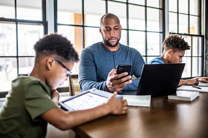 Father and two young sons sitting at a table with technology. The father is looking at his smartphone with his laptop open in front of him.