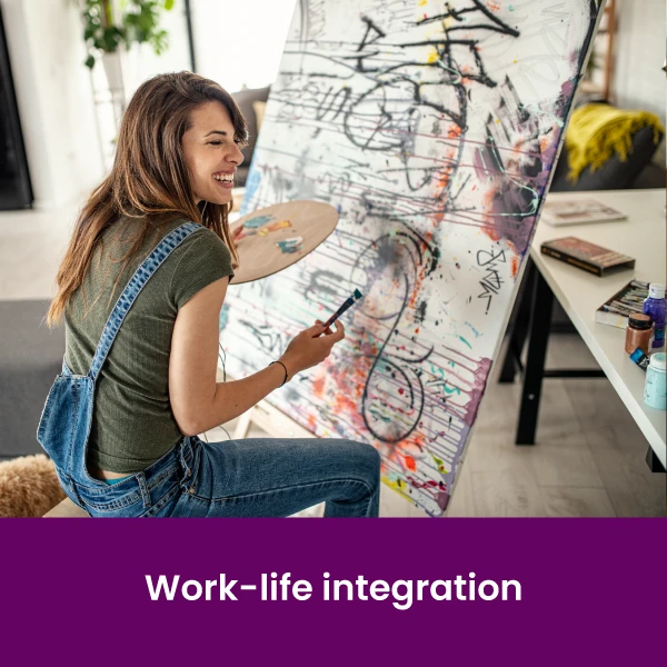 a smiling woman holding an easel sitting in front of a large canvas and painting