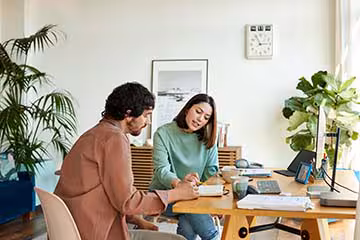 A man and woman are sitting at a desk and looking at a notebook together in a brightly lit home office.