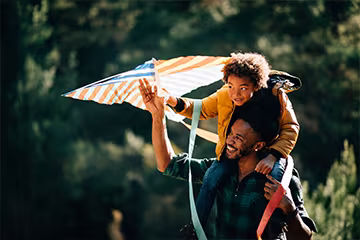 Boy sitting on his dad’s shoulders helps hold up a colorful kite