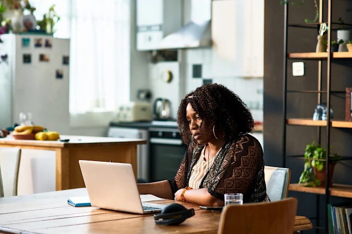 A woman sits at a counter in the kitchen facing a large window while looking at her phone.