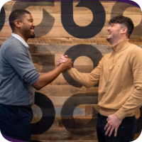 A young black man in a grey sweater and black pants shakes hands with a young white man in a tan sweater and black pants in front of a wooden wall with repeating lowercase a Ally logos on it.